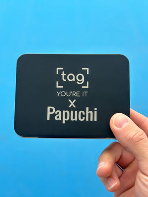 A hand holding a black square metal tin with rounded corners against a bright blue background. The tin is printed with the text 'tag YOU'RE IT x Papuchi' in white.